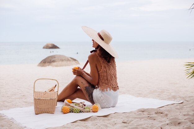 Giovane donna in forma abbronzata abbastanza caucasica in vestiti a maglia e cappello sulla spiaggia