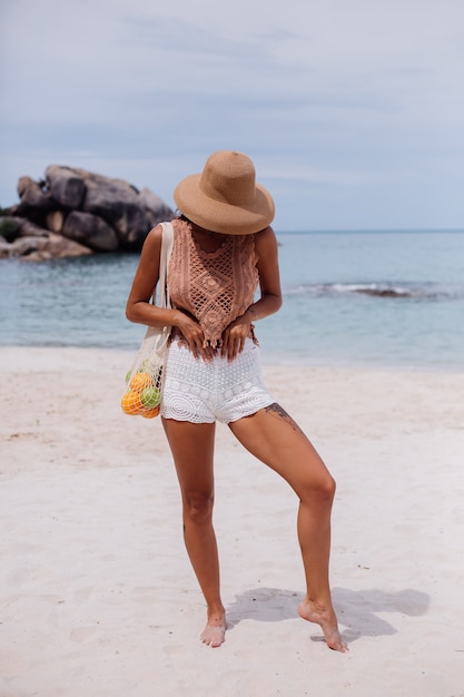 Giovane donna in forma abbronzata abbastanza caucasica in vestiti lavorati a maglia sulla spiaggia tropicale che indossa il cappello di paglia che tiene il sacchetto di stringa riutilizzabile ecologia pieno di frutta mela arancia uva