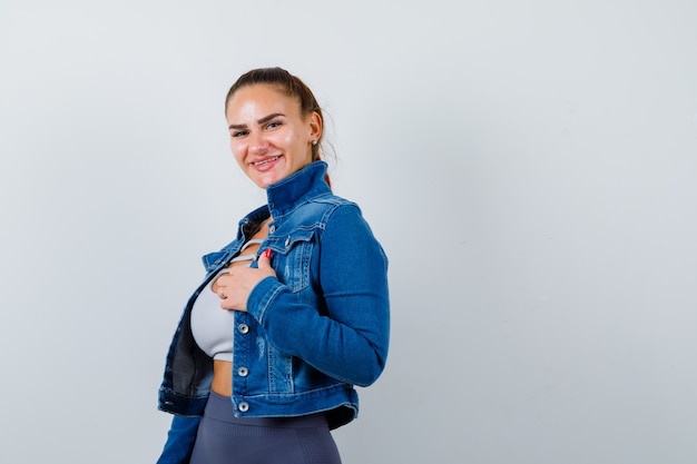 Giovane donna in forma in alto, giacca di jeans che tiene la mano sul petto mentre posa e sembra allegra, vista frontale.