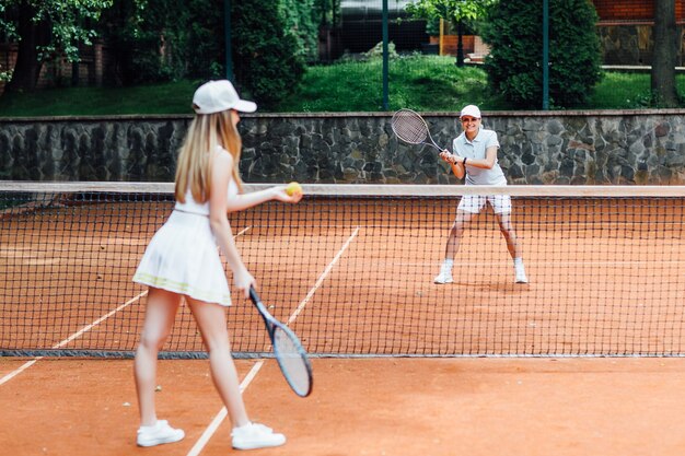 Giovane donna in forma in berretto e uniforme da tennis che serve pallina da tennis durante l'allenamento sul campo da tennis all'aperto.