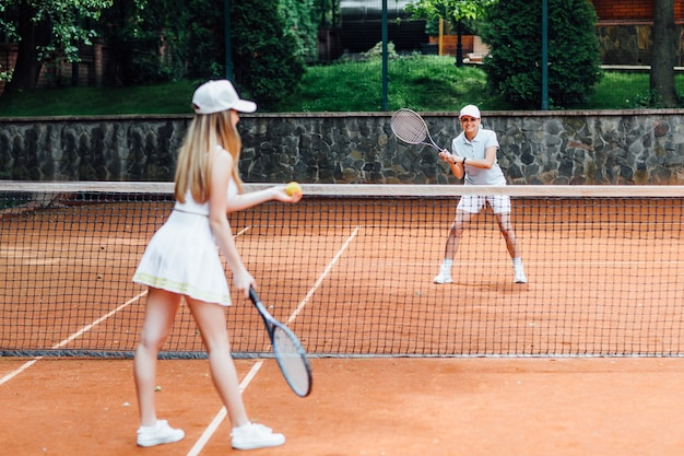 Giovane donna in forma in berretto e uniforme da tennis che serve pallina da tennis durante l'allenamento sul campo da tennis all'aperto.