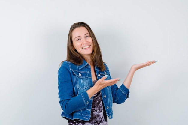 Giovane donna in giacca di jeans che mostra qualcosa sul muro bianco con spazio di copia e sembra allegra, vista frontale.