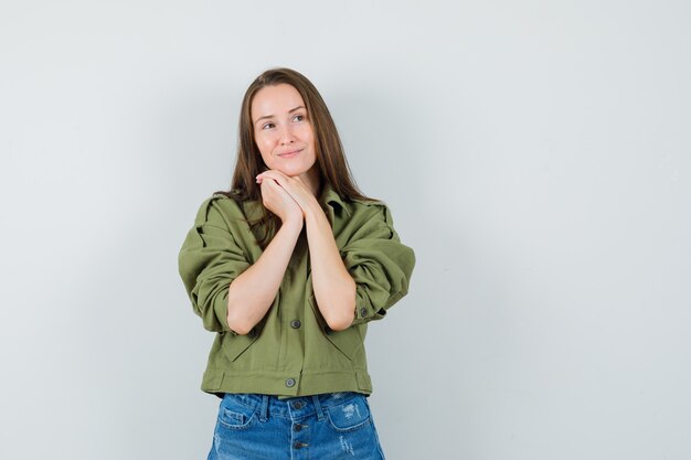 Giovane donna in giacca, pantaloncini appoggiando il mento sulle mani giunte e guardando sognante, vista frontale.