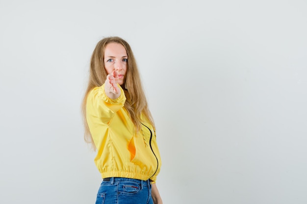 Giovane donna in giubbotto bomber giallo e jeans blu che punta alla telecamera e guardando serio, vista frontale.