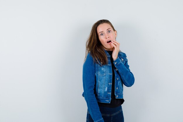 Giovane donna in jeans camicetta che tiene la mano vicino alla bocca aperta e sembra sorpresa, vista frontale.