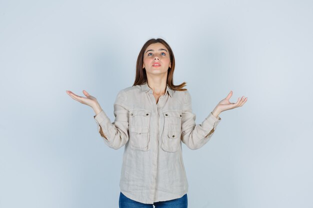 Giovane donna in jeans casual, allargando i palmi delle mani mentre guarda in alto e sembra meravigliata, vista frontale.