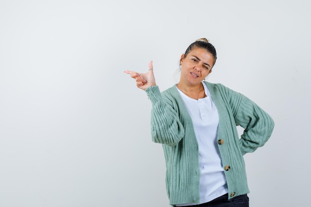 Giovane donna in maglietta bianca e cardigan verde menta che punta a sinistra con il dito indice, strizza l'occhio e sembra attraente