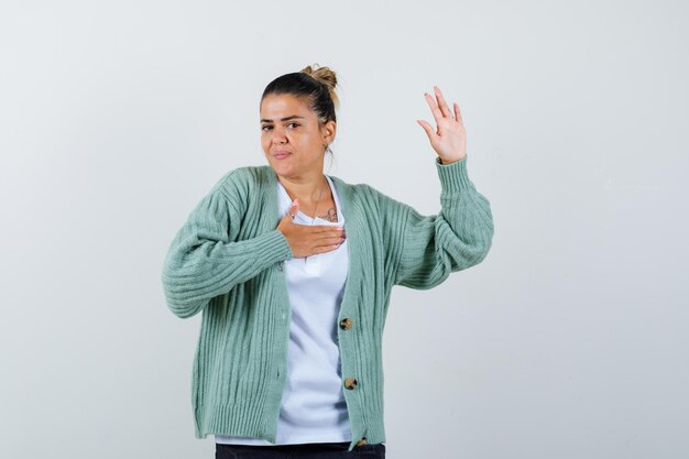 Giovane donna in maglietta bianca e cardigan verde menta che tiene la mano sul petto mentre alza la mano per salutare e sembra felice and