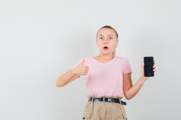 Giovane donna in maglietta e pantaloni che tiene il telefono cellulare, mostrando il pollice in su e guardando stupito