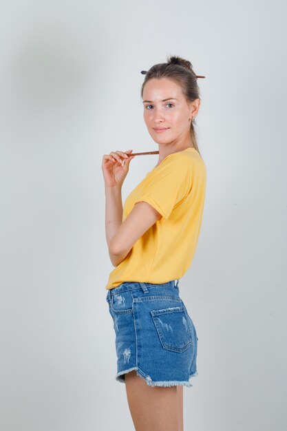 Giovane donna in maglietta gialla, pantaloncini di jeans che guarda l'obbiettivo con pennello da pittura e guardando allegro