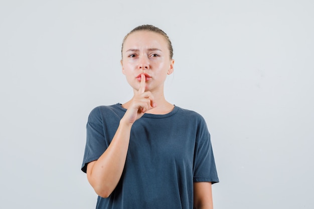 Giovane donna in maglietta grigia che mostra gesto di silenzio e guardando attento