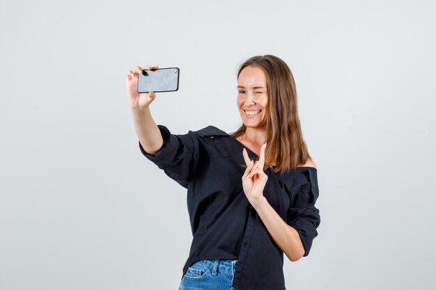 Giovane donna in maglietta, pantaloncini che mostra il segno v mentre si prende selfie e sembra felice