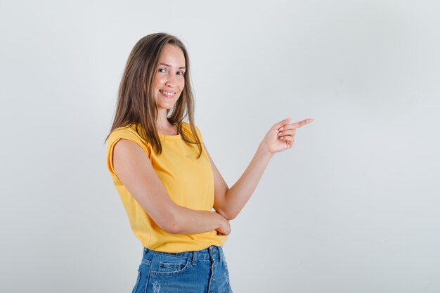 Giovane donna in maglietta, pantaloncini puntando il dito a lato e guardando allegra