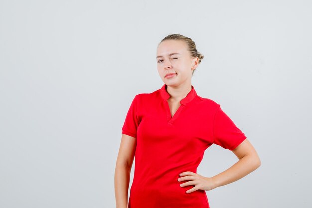 Giovane donna in maglietta rossa ammiccante con gli occhi e tenendo la mano sulla vita