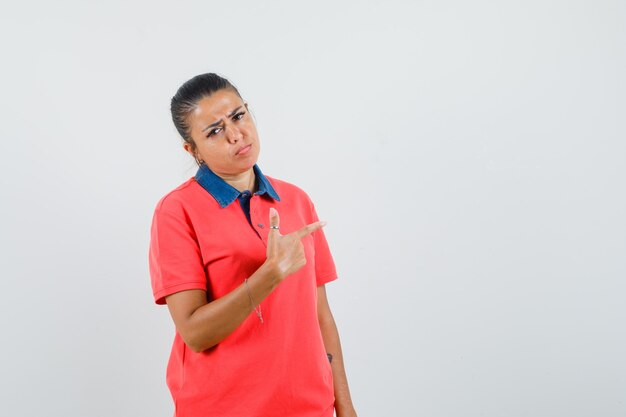 Giovane donna in maglietta rossa che punta a destra con il dito indice, guance gonfie e triste, vista frontale.