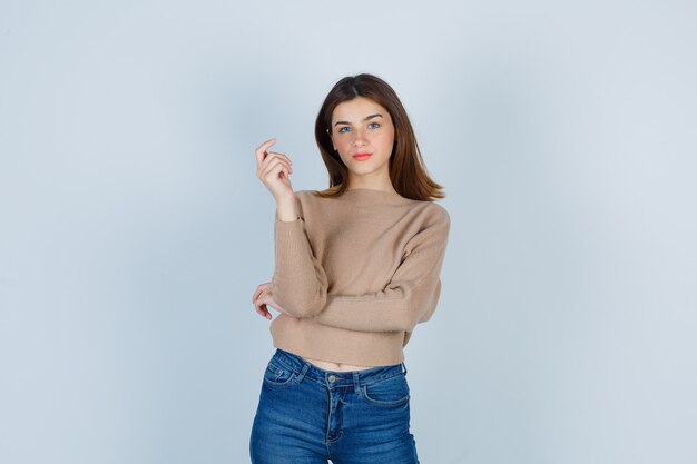 Giovane donna in maglione beige, jeans in posa mentre guarda davanti e sembra sicura, vista frontale.