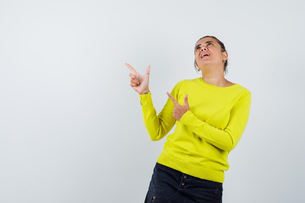 Giovane donna in maglione giallo e pantaloni neri che punta verso l'alto, guarda in alto e sembra felice