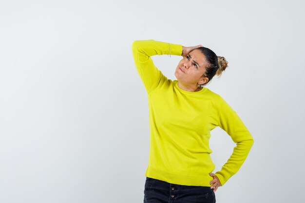 Giovane donna in maglione giallo e pantaloni neri in piedi in posa di pensiero e guardando pensierosa