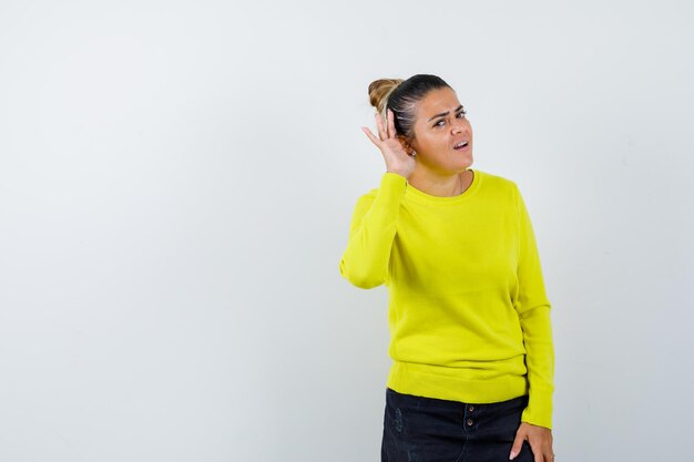 Giovane donna in maglione, gonna di jeans che tiene la mano dietro l'orecchio e sembra meravigliata