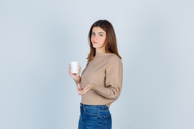 Giovane donna in maglione, jeans che tiene un bicchiere di carta, dispiaciuta per la domanda stupida e con l'aria malinconica.