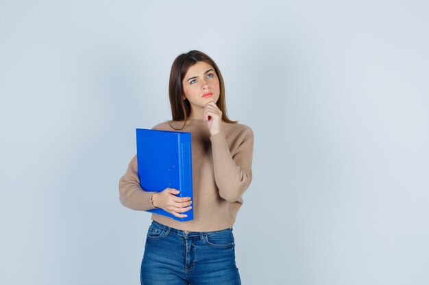 Giovane donna in maglione, jeans con la mano sul mento, tenendo la cartella e guardando malinconico, vista frontale.