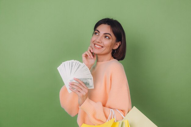 Giovane donna in maglione pesca casual isolato su parete verde oliva che tiene un ventilatore di 100 banconote da un dollaro soldi e borse della spesa pensando positivo sorridente copia spazio