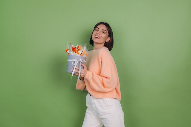 Giovane donna in maglione pesca casual isolato sul muro verde oliva tenere arancione fioriera bianca composizione di fiori di cotone gypsophila grano e lagurus per un regalo felice stupito sorpreso