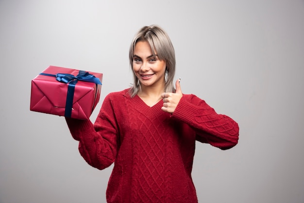 Giovane donna in maglione rosso che tiene il regalo di Natale e dà i pollici in su.