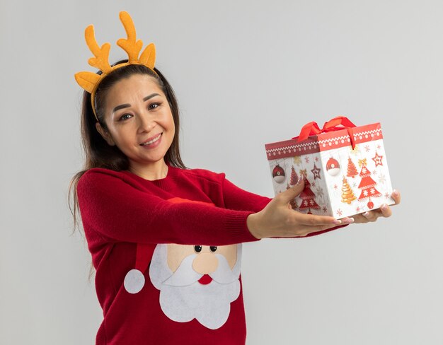 Giovane donna in maglione rosso di natale che indossa l'orlo divertente con le corna dei cervi che tengono il regalo di natale che sembra sorridere allegramente felice e positivo