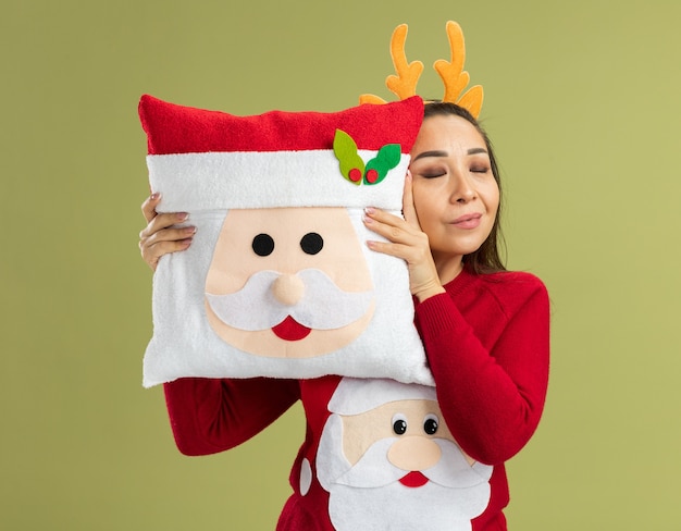 Giovane donna in maglione rosso di natale che indossa un bordo divertente con corna di cervo che tengono cuscino di natale felice e positivo con gli occhi chiusi in piedi sul muro verde