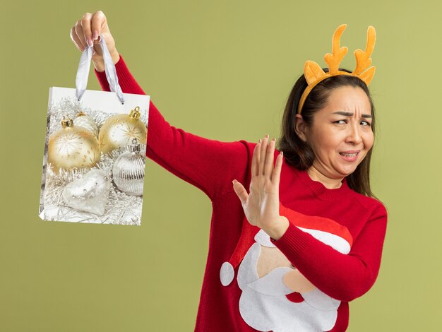 Giovane donna in maglione rosso di Natale che indossa un bordo divertente con corna di cervo che tiene un sacchetto di carta con un regalo di Natale guardandolo con dispiacere che tiene la mano in piedi sul muro verde