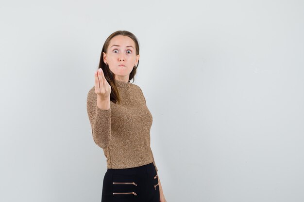 Giovane donna in maglione scivolato oro e pantaloni neri che mostra gesto italiano e sembra sorpresa, vista frontale.