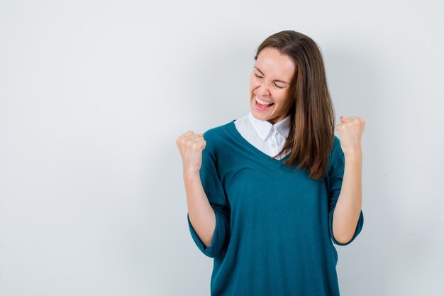 Giovane donna in maglione sopra la camicia bianca che mostra il gesto del vincitore e sembra felice, vista frontale.