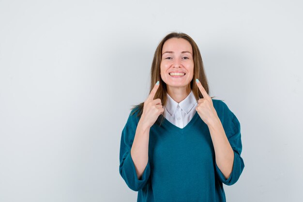 Giovane donna in maglione sopra la camicia che punta ai suoi denti e sembra gioiosa, vista frontale.