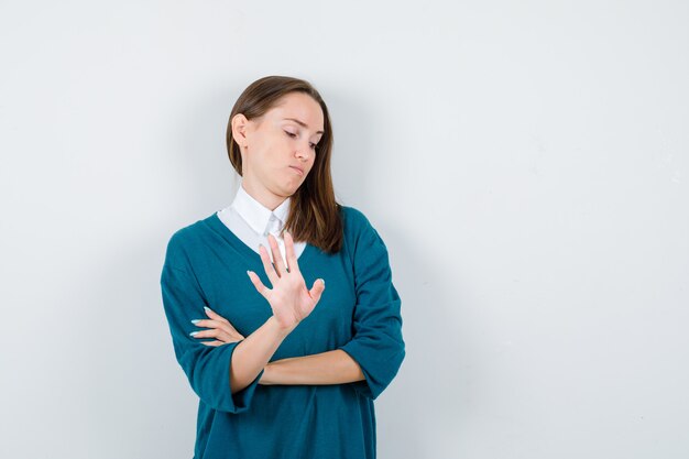 Giovane donna in maglione sopra una camicia bianca che mostra il gesto di arresto e sembra riluttante, vista frontale.