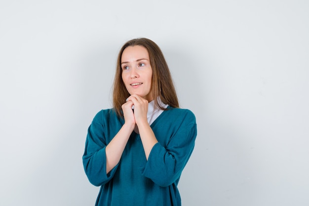 Giovane donna in maglione su camicia bianca che guarda lontano con il mento appoggiato sulle mani giunte e dall'aspetto sognante, vista frontale.