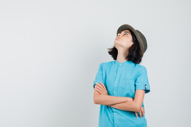 Giovane donna in piedi con le braccia incrociate, guardando il soffitto in camicia blu, cappello