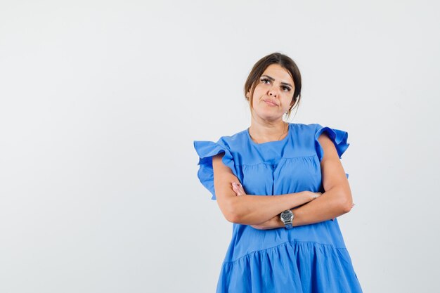 Giovane donna in piedi con le braccia incrociate in abito blu e guardando pensieroso