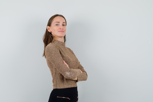 Giovane donna in piedi con le braccia incrociate in camicetta dorata e guardando fiducioso, vista frontale. spazio per il testo