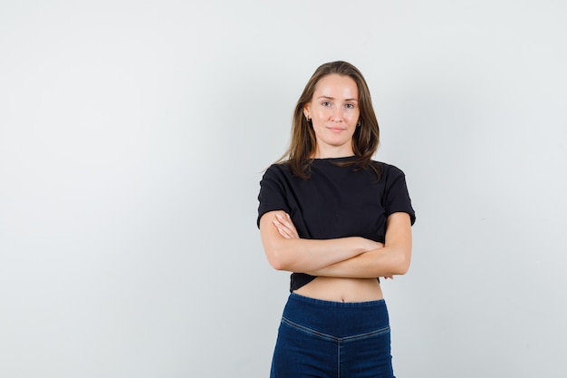 Giovane donna in piedi con le braccia incrociate in camicetta nera, pantaloni e guardando fiducioso