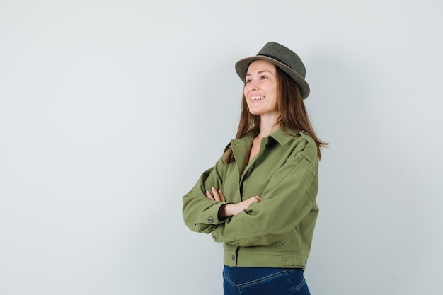 Giovane donna in piedi con le braccia incrociate in giacca pantaloni cappello e guardando allegra