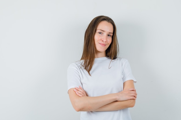 Giovane donna in piedi con le braccia incrociate in maglietta bianca e guardando fiducioso