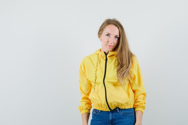 Giovane donna in piedi dritto e posa alla macchina fotografica in bomber giallo e jeans blu e guardando affascinante, vista frontale.