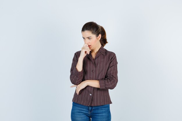 Giovane donna in piedi in posa di pensiero, con la mano davanti alla bocca in camicia a righe, jeans e guardando pensieroso, vista frontale.