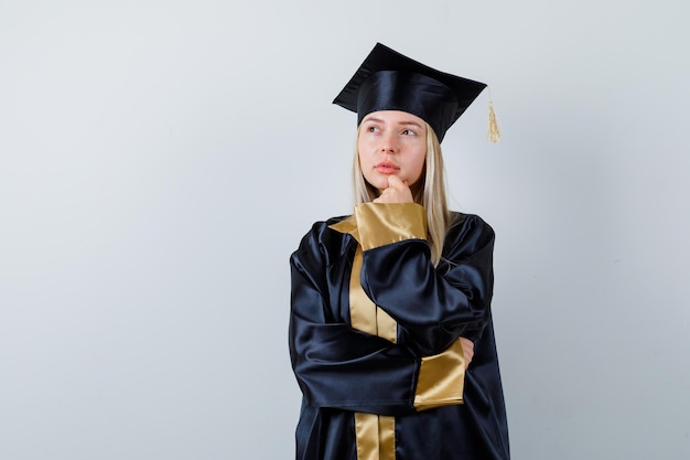 Giovane donna in piedi in posa di pensiero in abito accademico e guardando pensierosa