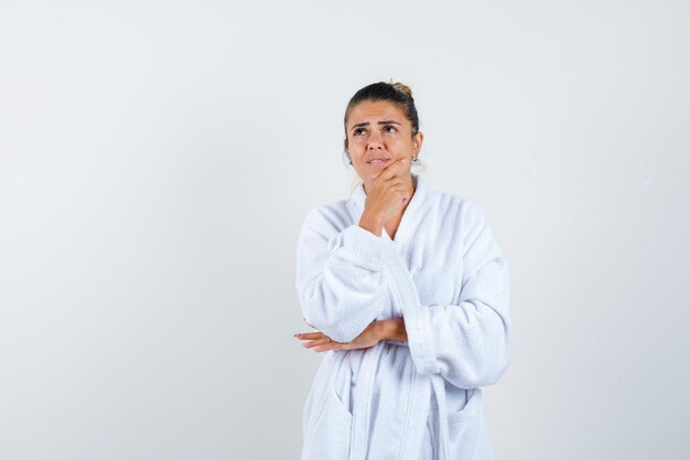 Giovane donna in piedi in posa di pensiero in accappatoio e guardando pensierosa