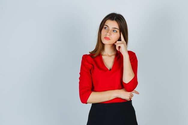 Giovane donna in piedi nel pensiero posa in camicetta rossa, gonna e guardando pensieroso