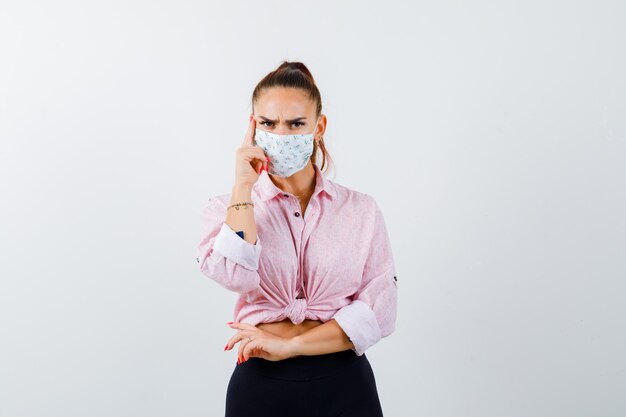 Giovane donna in piedi nel pensiero posa in camicia, pantaloni, maschera e guardando serio, vista frontale.