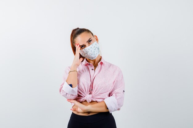 Giovane donna in piedi nel pensiero posa in camicia, pantaloni, mascherina medica e guardando pensieroso. vista frontale.