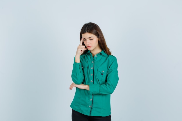 Giovane donna in piedi nella posa di pensiero in camicia verde e guardando turbato. vista frontale.